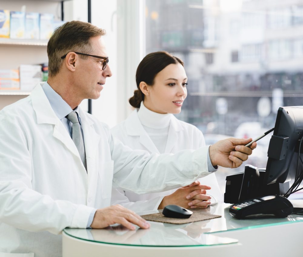 pharmacist pointing showing male colleague medication prices prescriptions on computer screen